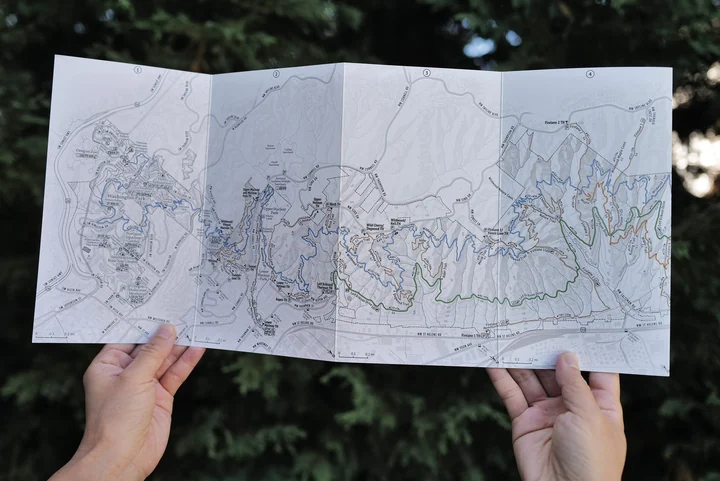Two hands hold the Pocket Adventure Map fully unfolded in front of Forest Park greenery.